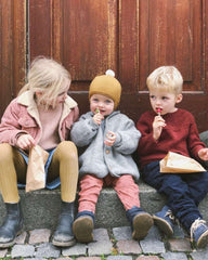 Tre siddende børn. Barnet i midten er iført en gul Alberts Pilothue.