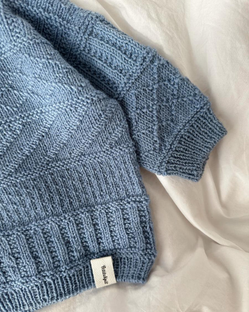 Lyseblå Storm Sweater Baby liggende på sengetøj