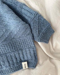 Lyseblå Storm Sweater Baby liggende på sengetøj